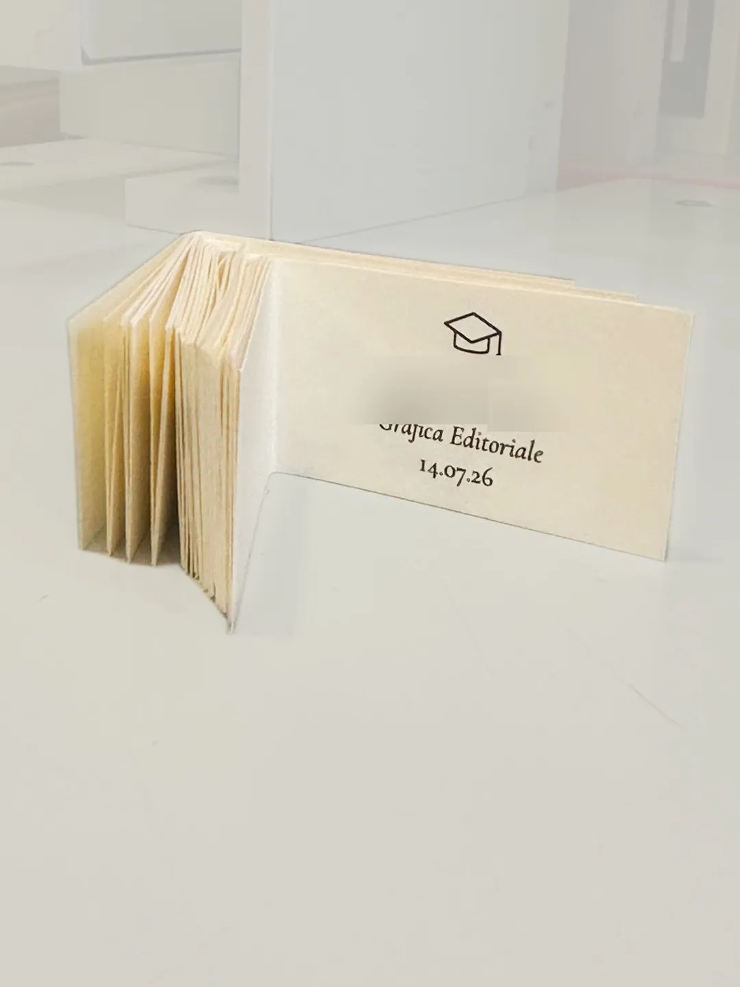 Graduation favour cards printed by GUUG: a fanned stack of folded ivory cards with an academic cap icon, degree programme and ceremony date (name blurred for privacy)