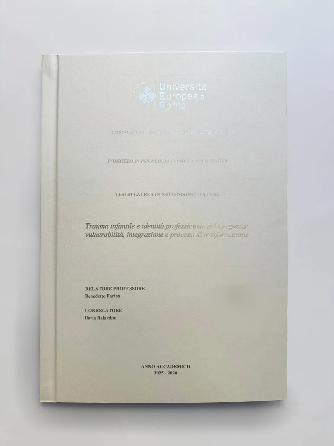 Tesi Università Europea di Roma rilegata da GUUG: copertina rigida bianco perla con logo UER e scritte a caldo argento, corso di laurea in Psicologia (nome del laureando sfocato per riservatezza)